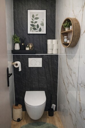 Un baño moderno con inodoro blanco, pared de mármol negro, una obra de arte con hojas enmarcada, repisa con artículos de aseo y una cesta tejida con una planta.