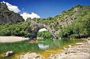 Une arche naturelle formée par la roche, enjambant une rivière aux eaux claires, et entourée de verdure et d’un paysage rocheux.