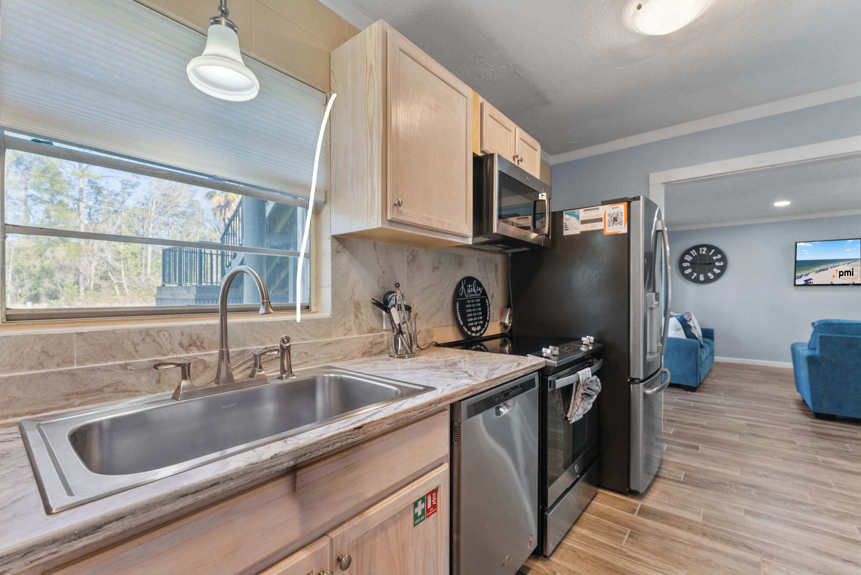 A modern kitchen with a sink, dishwasher, microwave, and refrigerator. There is a window providing natural light and a clock on the wall.