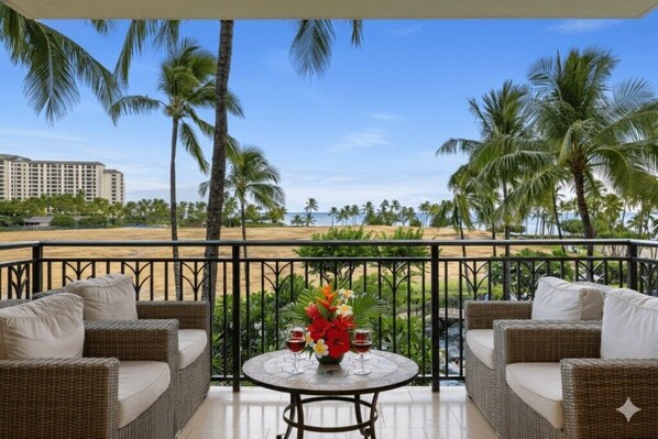 Een balkon met rieten meubels en een tafel met bloemen en wijnglazen, met uitzicht op een strand met palmbomen en een hotel op de achtergrond.
