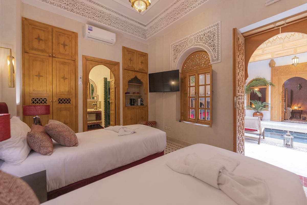 A hotel room with two beds, a TV, wooden cabinets, and a view of an outdoor pool area.