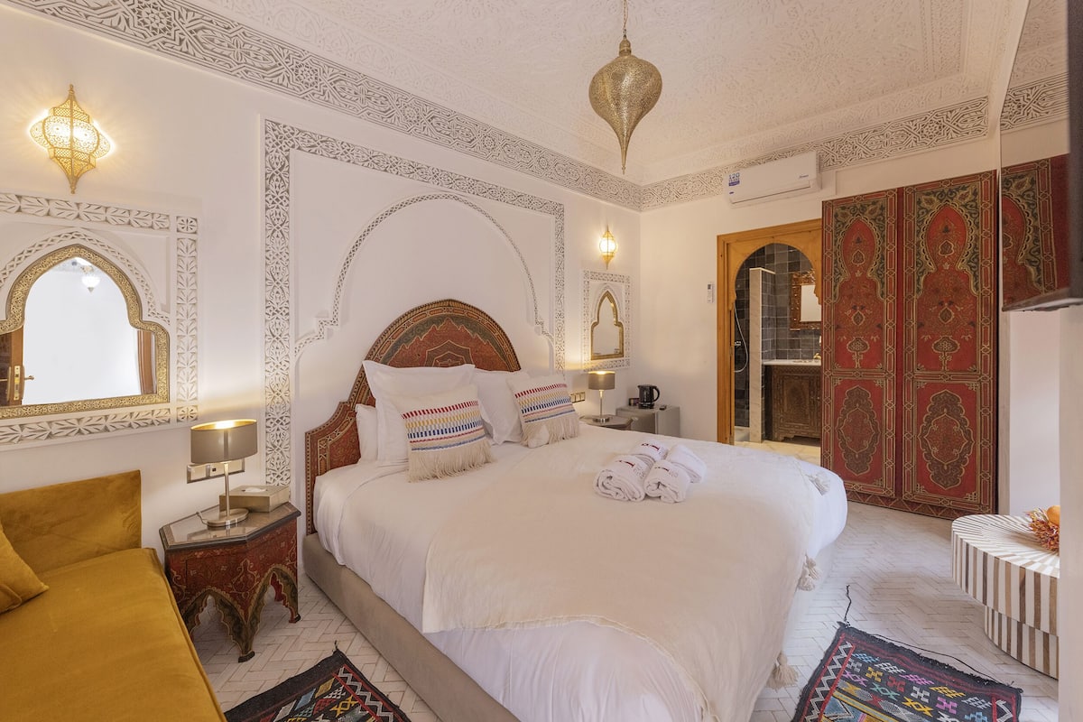 A hotel room with a large bed, a nightstand, a lamp, a mirror, and a wardrobe.