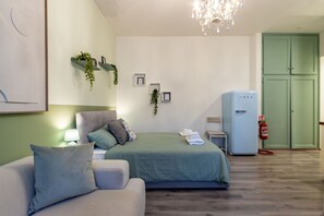 Un dormitorio con cama, sofá, candelabro, estante para plantas en la pared, una mesita, refrigerador y una puerta verde.