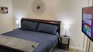 Ein Schlafzimmer mit einem Bett, zwei Nachttischlampen, einer Wanddekoration, einem Fernseher, der Streaming-Dienste anzeigt, und einem kleinen Tisch mit einer Fernbedienung.