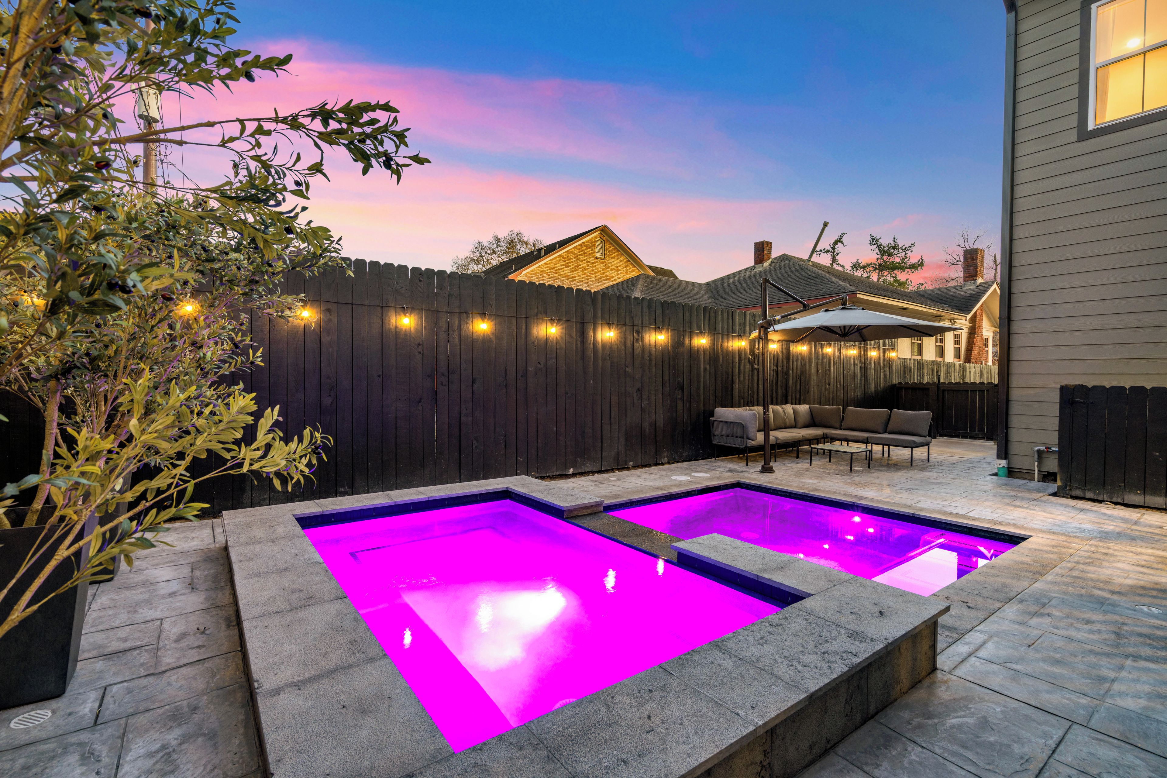 A backyard with two illuminated swimming pools, a wooden fence, and a patio with outdoor furniture.