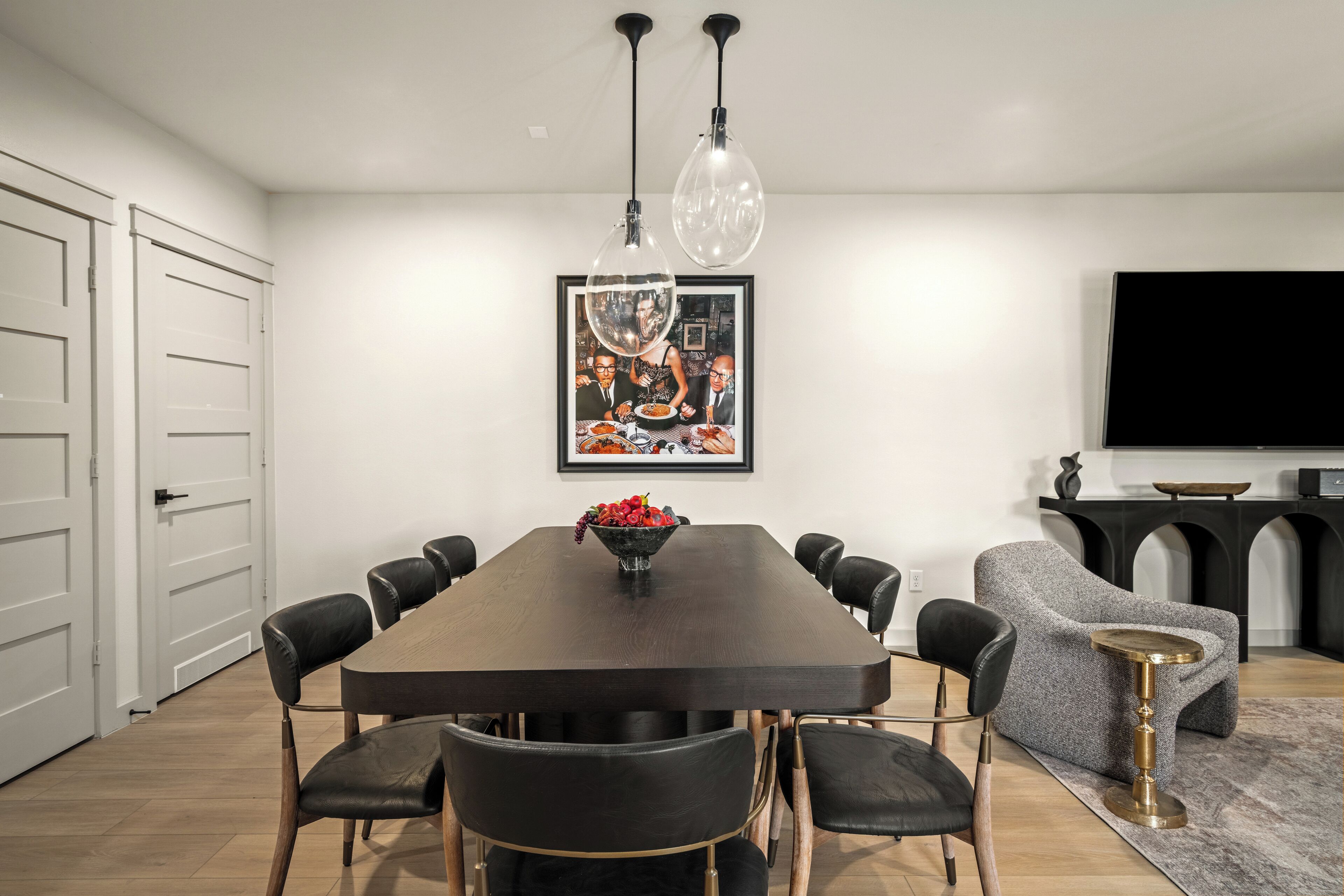 A modern dining area with a dark wooden table, black chairs, a fruit bowl, and a framed picture on the wall.