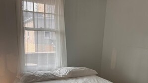 Une petite chambre avec un lit simple, recouverte d’une housse de couette blanche unie, dotée d’un rideau transparent et d’un plancher en bois.