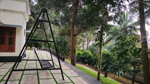A swing set in a lush garden area with a paved walkway and a building with a white facade and wooden window frames. - Elove Resort Private Hideaway on 6 Serene Acres (Thamarassery)