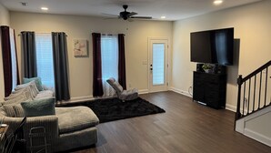 Ein Wohnzimmer mit einer Couch, einem Sessel, einem schwarzen Teppich, einem Deckenventilator und einem an der Wand befestigten Fernseher.