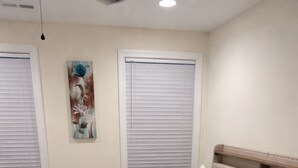 Ein Schlafzimmer mit einem Bett, zwei Fenstern mit Jalousien, einem Bild an der Wand und einer Deckenleuchte.