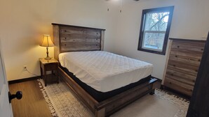 Een slaapkamer met een houten hoofdbord, een bed met een witte deken, een nachtkastje met een lamp en een raam met uitzicht op bomen.