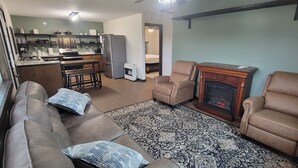 Een woonkamer met een bank, twee fauteuils, een open haard en een keuken met een koelkast en aanrecht.