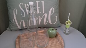 Ein runder Tisch mit einer Ablage, auf der zwei Gläser, eine Flasche und ein Apfel stehen. Dahinter ein Kissen mit der Aufschrift 'home sweet home'.