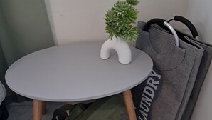 Ein kleiner runder Tisch mit einer weißen Vase, darin eine grüne Pflanze, einer grauen Wäschetasche und einem grauen Korb, unter dem ein grünes Handtuch liegt.