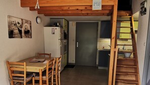 A compact kitchen with a refrigerator, a dining table with chairs, and a wooden staircase leading to an upper level. - Practical house, pets welcome - 6 guests (La Tranche-sur-Mer)