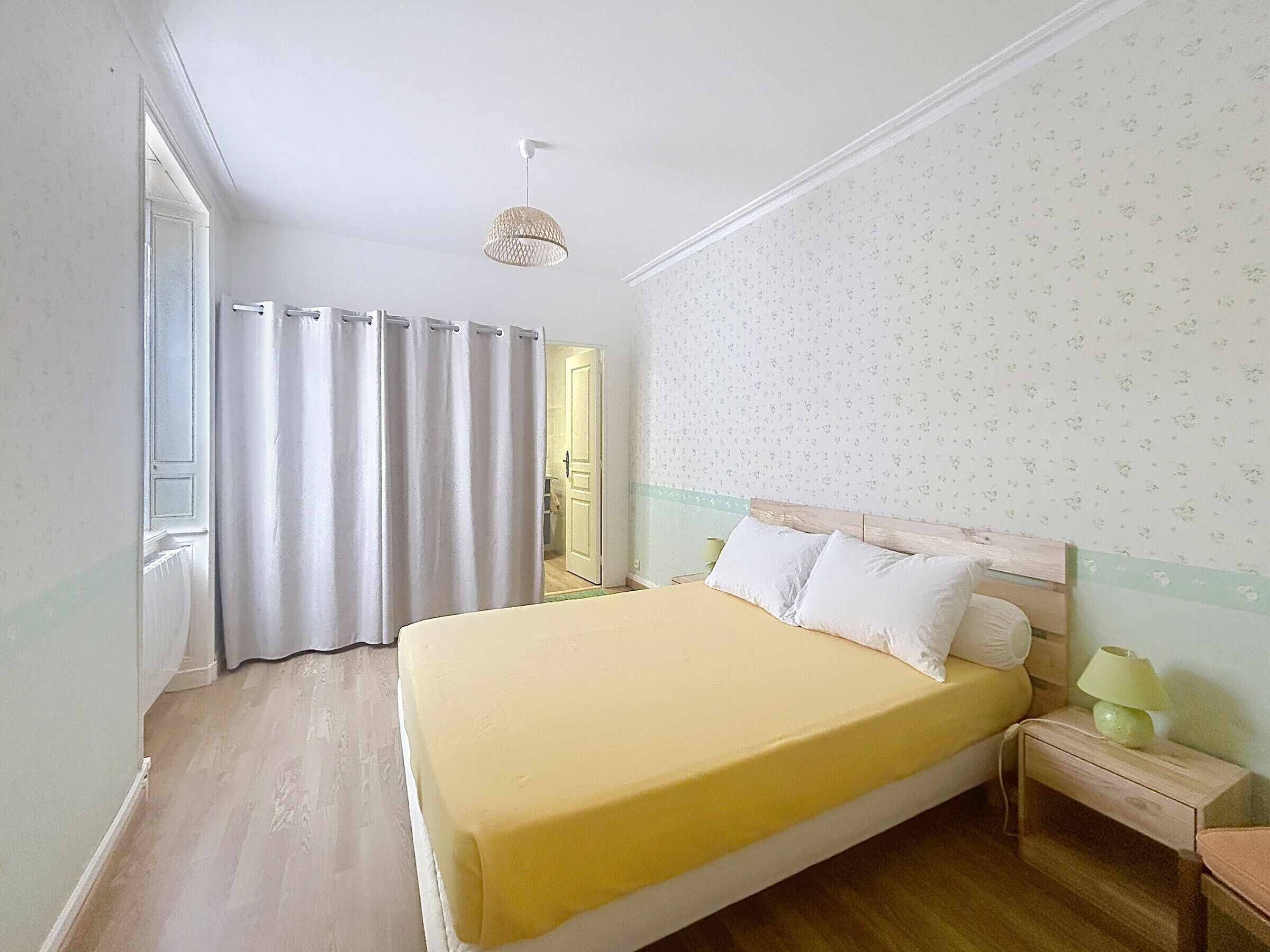 A bedroom with a bed, curtains, a nightstand, a lamp, and a door.