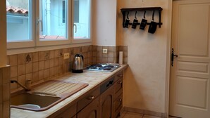 Cozinha compacta com fogão a gás, chaleira e pia. Há uma janela acima da pia e uma porta à direita.