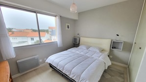 Een slaapkamer met een groot raam, een bed, een nachtkastje, een radiator en een klein plankje.