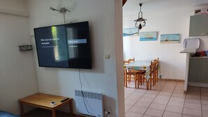 A room with a bed, a TV mounted on the wall, a small table, a radiator, a dining table with chairs, and a kitchen area. - Comfortable villa, parking, pets welcome - 6 guests (Gruissan)