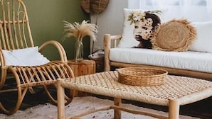 Een rieten stoel en een klein tafeltje met een gevlochten mand, een kussen met een luipaardprint kussen en een decoratieve bladwandhanger.
