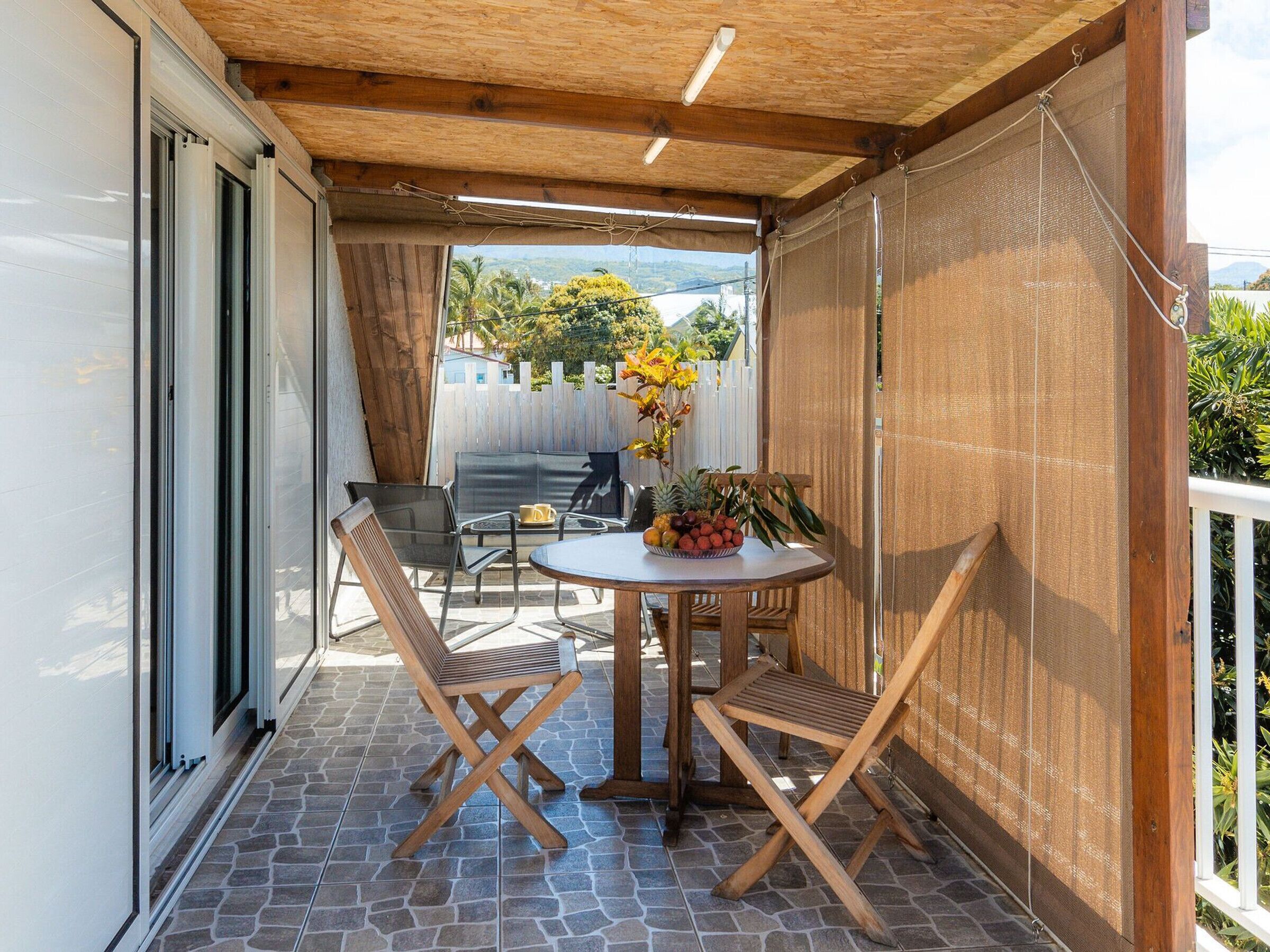 A covered patio with a table and two chairs, a white fence, and a view of a residential area.