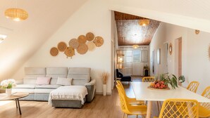 A cozy living room with a sofa, a dining table with yellow chairs, and a kitchen in the background.