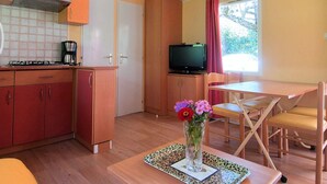 Uma cozinha compacta com fogão a gás, cafeteira e televisão sobre um armário. Possui um espaço de refeições com mesa e cadeiras, e um vaso com flores sobre a mesa.