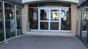 Una entrada de edificio con fachada de vidrio, el número '54' sobre las puertas y un escalón pequeño que lleva a la entrada.