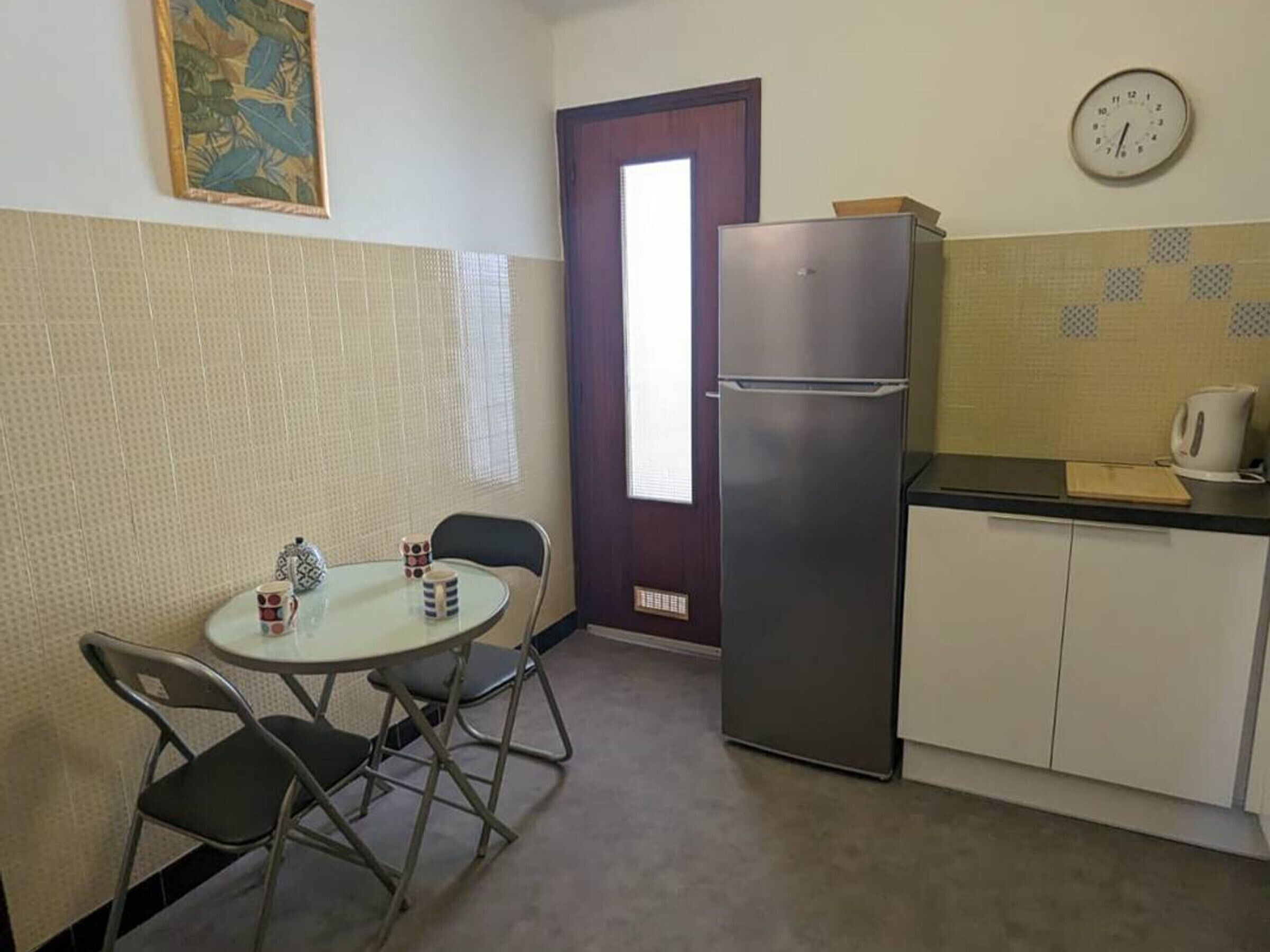 A compact kitchen with a refrigerator, a small dining table with chairs, a clock on the wall, and a window with blinds.