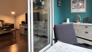 En moderne stue med en glasdør der fører ud til en balkon, en sofa, et lille bord og et skrivebord med en lampe.