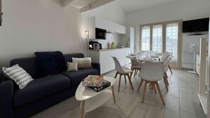 Een moderne woonkamer met een donkere bank, een bijzettafeltje en een eethoek met witte stoelen en een kleine tafel.
