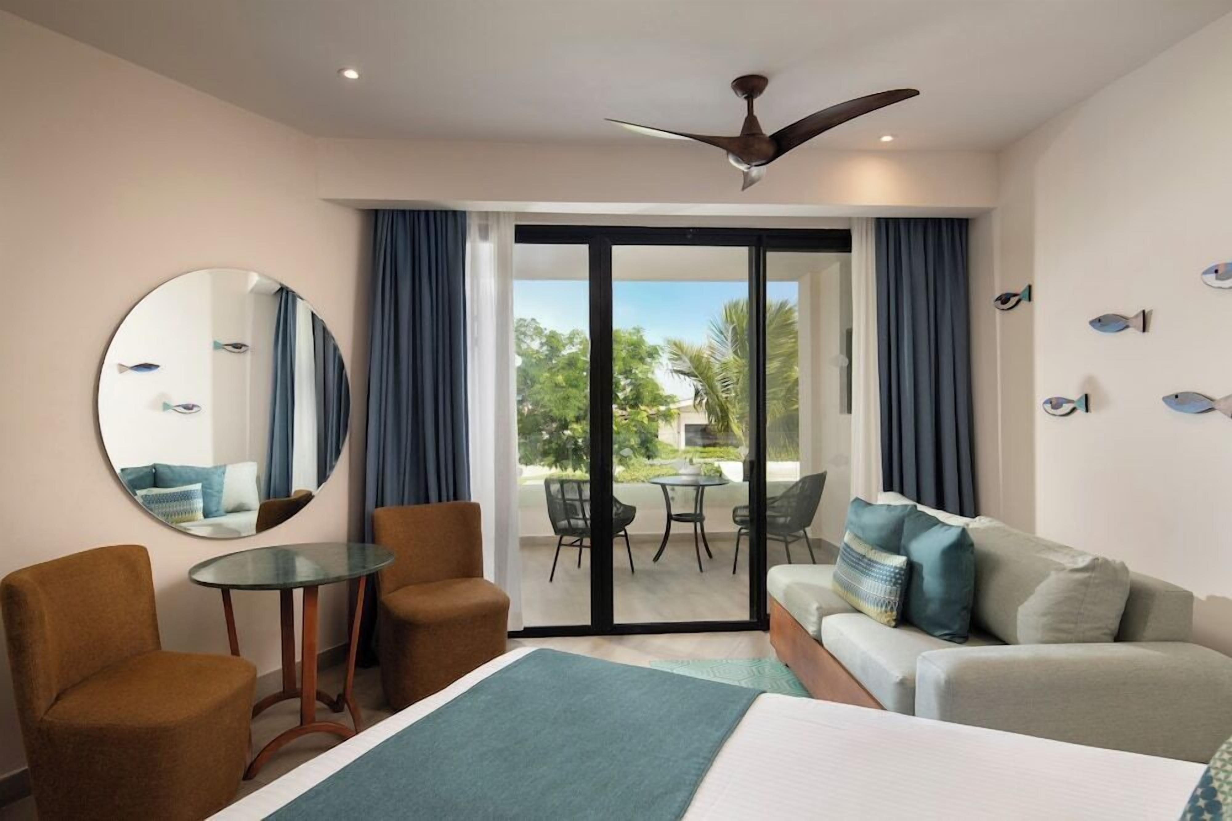 Ein Hotelzimmer mit einem Bett, zwei Sesseln, einem runden Spiegel, einem kleinen Tisch und einem Balkon mit Blick auf Grünflächen und ein Gebäude.