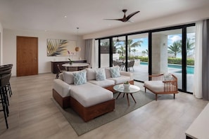 A modern living room with a sofa, a coffee table, and a chair, a dining area with a table and chairs, and a large sliding glass door leading to a pool area. - Dreams Macao Beach Punta Cana (Punta Cana)