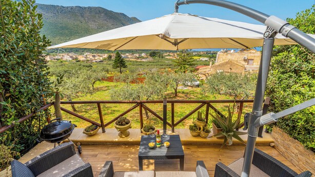 Un patio al aire libre con mobiliario de mimbre, una mesa con bebidas y un amplio paraguas que da sombra.