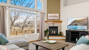 A living room with a fireplace, a television, and a large window overlooking a landscape. - Cabin-style Group Retreat Near Rmnp, Lakes & Golf (Loveland)