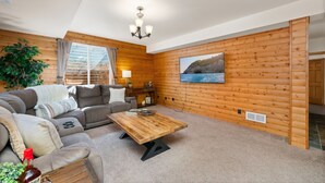 A cozy living room with a wooden wall, a large window, a sofa, a coffee table, and a painting of a coastal scene. - Cabin-style Group Retreat Near Rmnp, Lakes & Golf (Loveland)