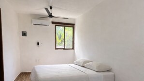 A bedroom with a bed, a ceiling fan, an air conditioning unit, and a window with a view of greenery.