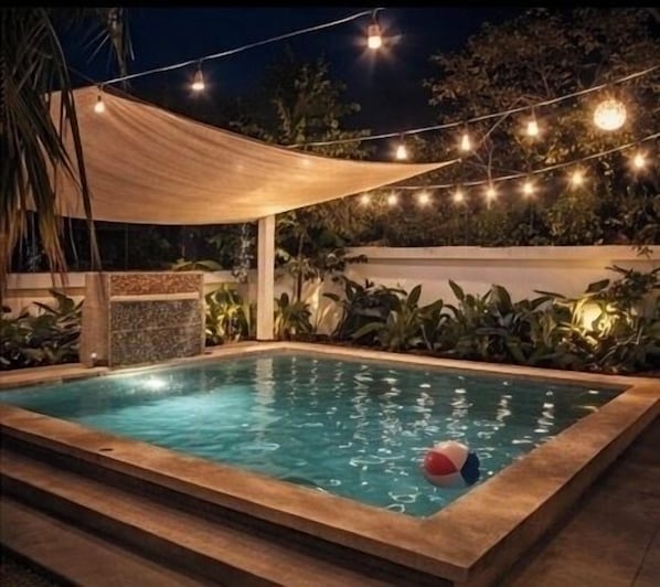 A rectangular swimming pool with a wooden deck, surrounded by plants and string lights.