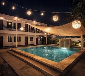 Ein Schwimmbad mit Holzterrasse, Lichterketten und einem Gebäude mit mehreren Balkonen.