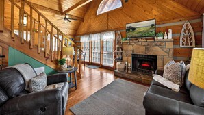 L’intérieur chaleureux d’un chalet avec une cheminée en pierre, un plafond en bois et un escalier menant à un niveau supérieur.