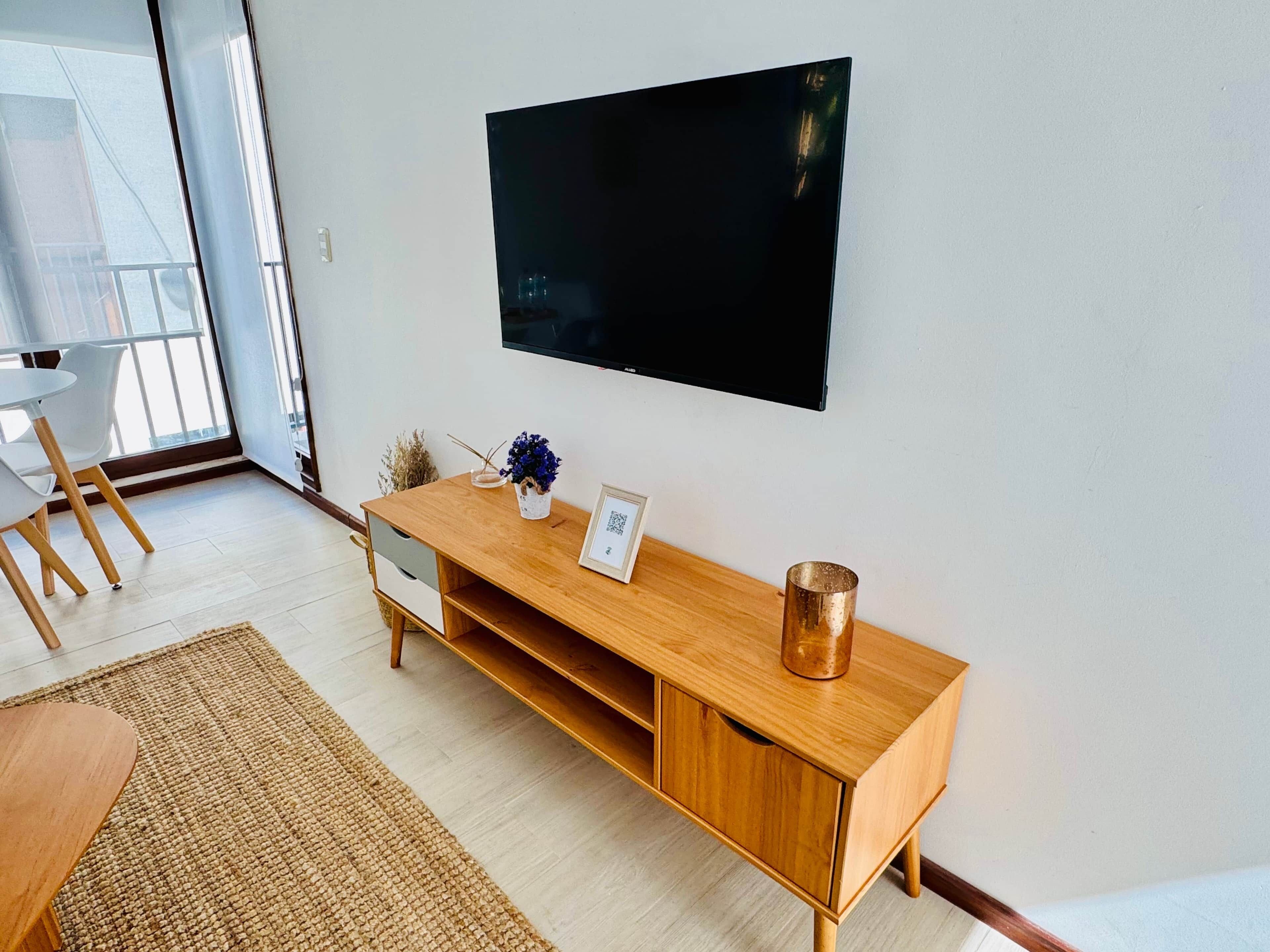 A modern living room with a wooden TV stand, a flat-screen TV mounted on the wall, a small table with chairs, and a large window providing natural light.