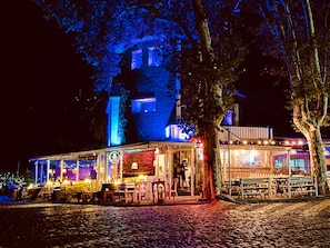 Edificio histórico con fachada de piedra, iluminado con luces azules y rojas, que cuenta con mesas al aire libre y una entrada visible.