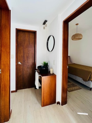 Une pièce avec une porte en bois, un miroir rond, un lavabo avec un receveur noir, une petite plante et un lit recouvert d’une couverture marron.