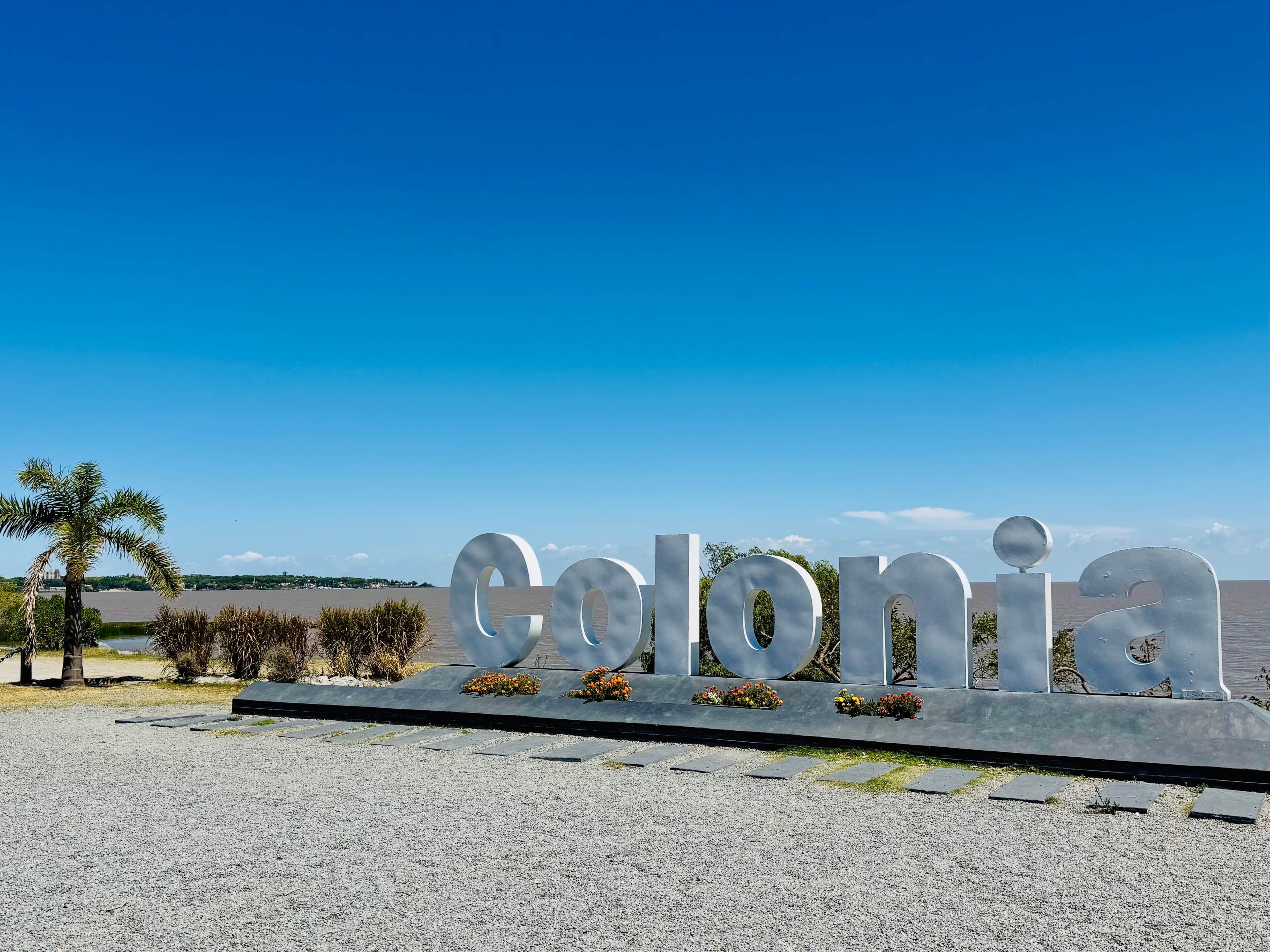 Un grande cartello metallico con la scritta "Colonia", sullo sfondo un cielo azzurro e un mare calmo.