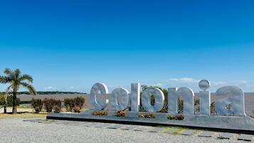 Un grande cartello metallico con la scritta "Colonia", sullo sfondo un cielo azzurro e un mare calmo.