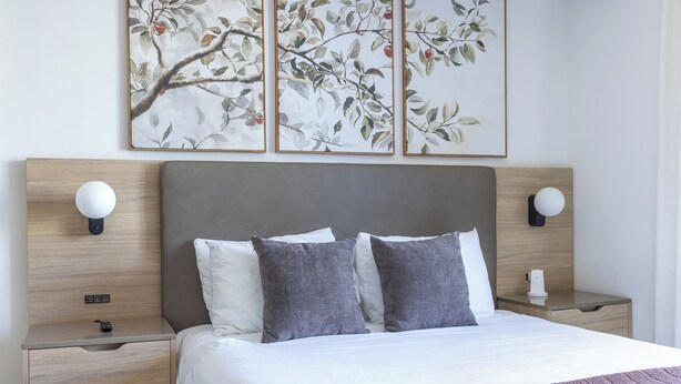 Una cama bien hecha con ropa de cama blanca y gris, un cabecero de madera y dos mesitas de noche con lámparas de pared.