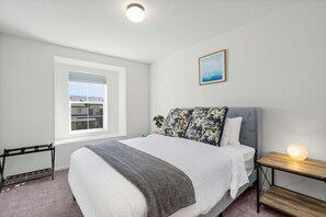 Een slaapkamer met een groot bed, een nachtkastje met een lamp, een raam met zonwering en een ingelijst schilderij aan de muur.