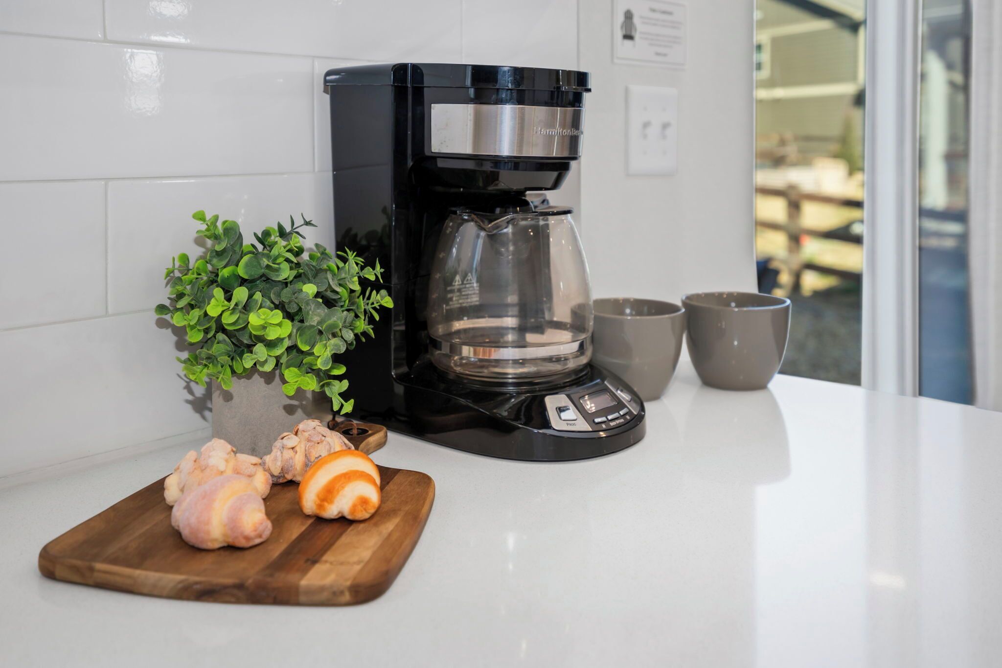A kitchen with a coffee maker, a potted plant, a cutting board with ginger, and two cups.