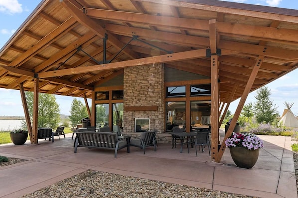 A covered patio with outdoor seating, a stone fireplace, and large windows.