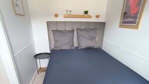 A bedroom with a bed, pillows, a bedside table, a chair, a shelf with decorative items, and a poster on the wall. - Cosy small holiday apartment for 4 persons (Tranekær)